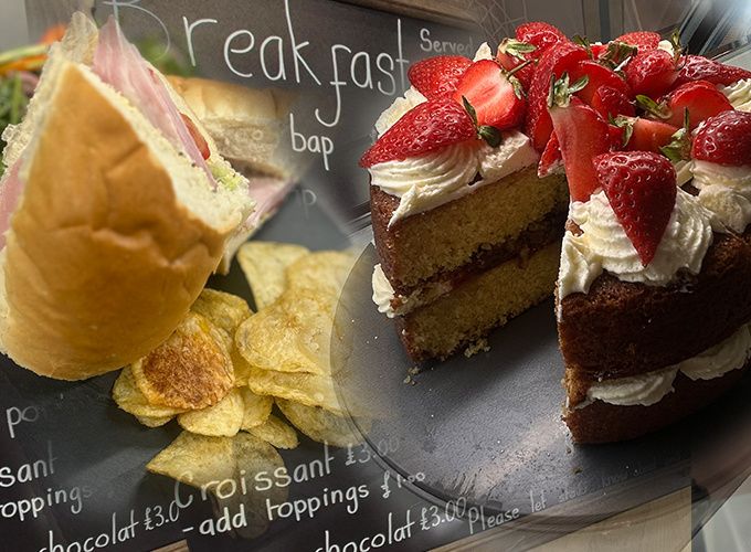 A filled roll with crisps and a strawberry sponge cake in front of the breakfast chalkboard, beside the espresso machine.