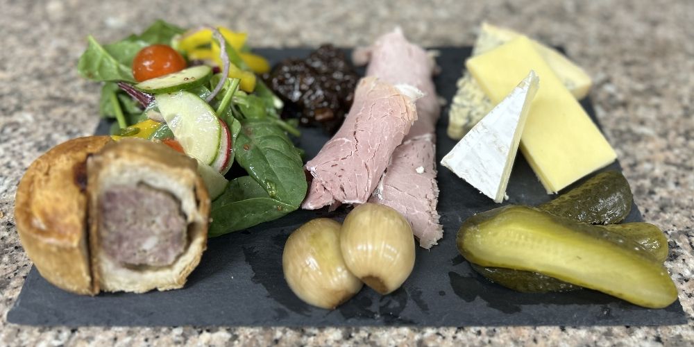 A ploughman's lunch on a slate board: pork pie, ham, two cheeses, pickled onions, gherkin and salad.